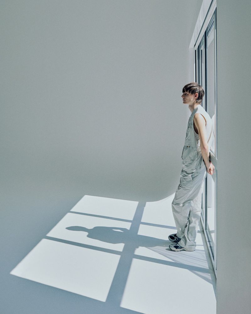 Young adult in a light blue jumpsuit stands near a window in a bright photo studio, casting a shadow on the floor.