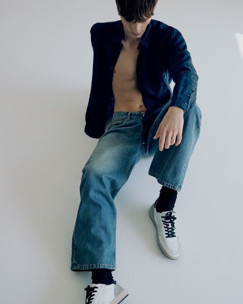 Male fashion model poses in an unbuttoned blue shirt and baggy jeans against a white backdrop, showcasing casual yet stylish apparel.