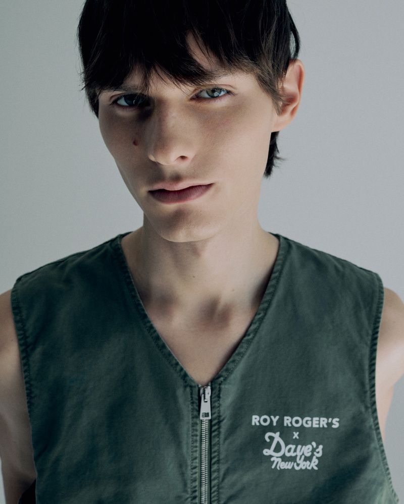 Close-up of a young adult male model posing in a sleeveless olive green vest with a zipper and a logo on the chest in a studio shot.