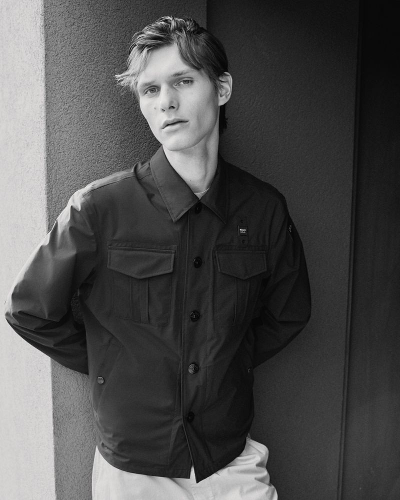 Black-and-white portrait of a young adult male wearing a dark collared jacket, standing against a textured gray wall, in a studio setting.