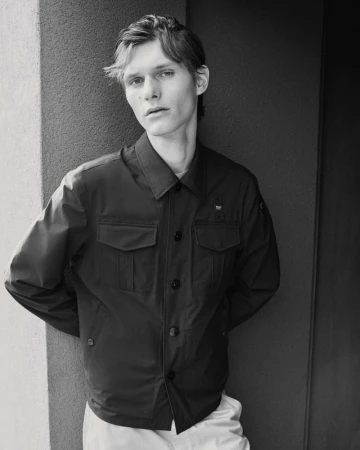 Black-and-white portrait of a young adult male wearing a dark collared jacket, standing against a textured gray wall, in a studio setting.