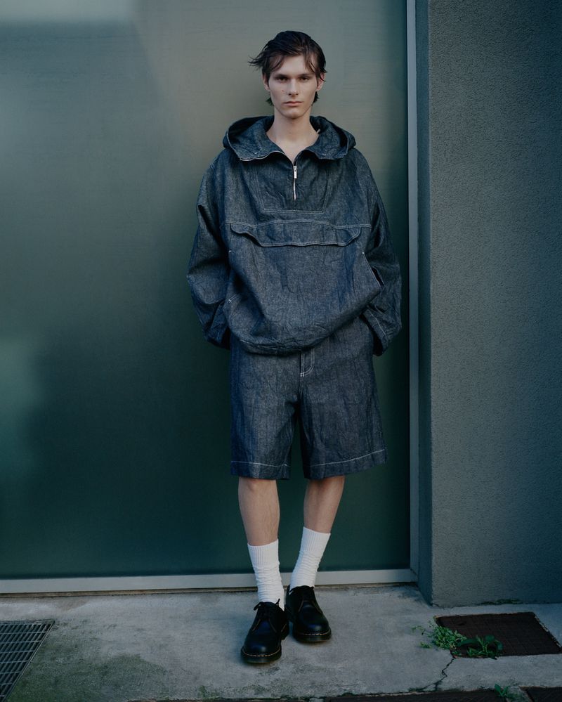 Adult male in anorak and shorts posing against a green wall in outdoor setting, wearing white socks and black leather shoes.