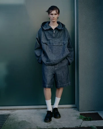 Adult male in anorak and shorts posing against a green wall in outdoor setting, wearing white socks and black leather shoes.