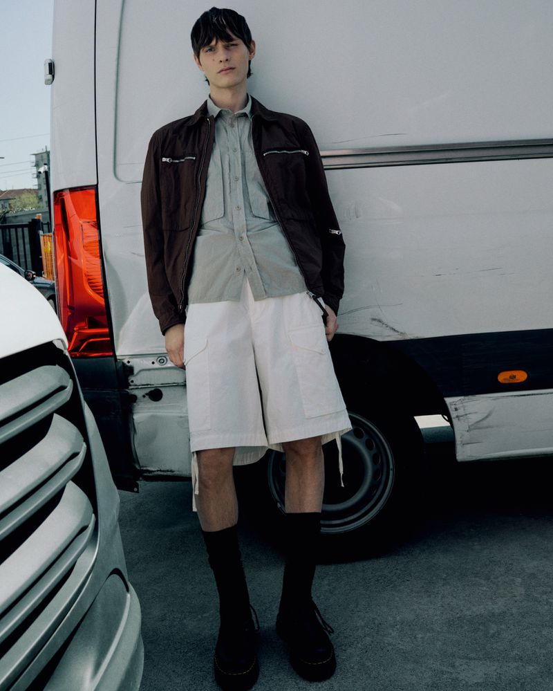 A stylish adult male model in a casual brown jacket, white shorts, and black socks poses against a white van outdoors.