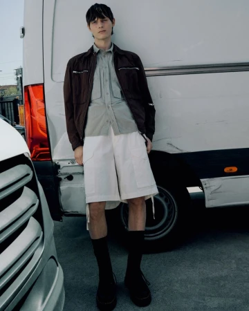 A stylish adult male model in a casual brown jacket, white shorts, and black socks poses against a white van outdoors.
