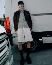Model leaning against a white van wearing a brown jacket, grey shirt, and white shorts