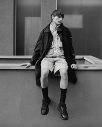 Monochrome fashion portrait of a male model seated on a ledge wearing a utility jacket, shorts, and dark socks with shoes.