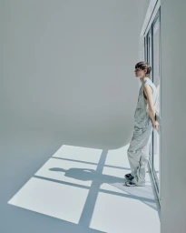 A young male model standing by a tall window wearing pale overalls in a bright studio space casting sharp shadows on the floor.