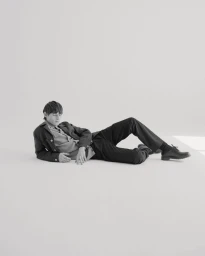 A man wearing a dark jacket and trousers lies on a white studio floor for an editorial portrait.
