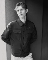 Monochrome portrait of a young male model leaning against a textured architectural wall wearing a dark utility jacket