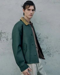 Young male model wearing a dark green jacket and tan trousers posing against a weathered pale wall for Esquire Italia.