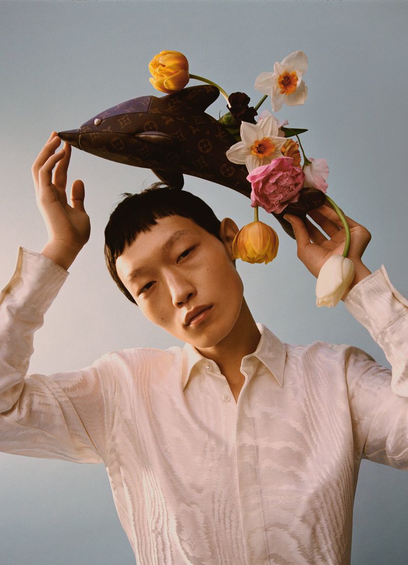 Asian male model poses with a Louis Vuitton dolphin purse adorned with tulips and daffodils against a light blue backdrop in a studio shot.