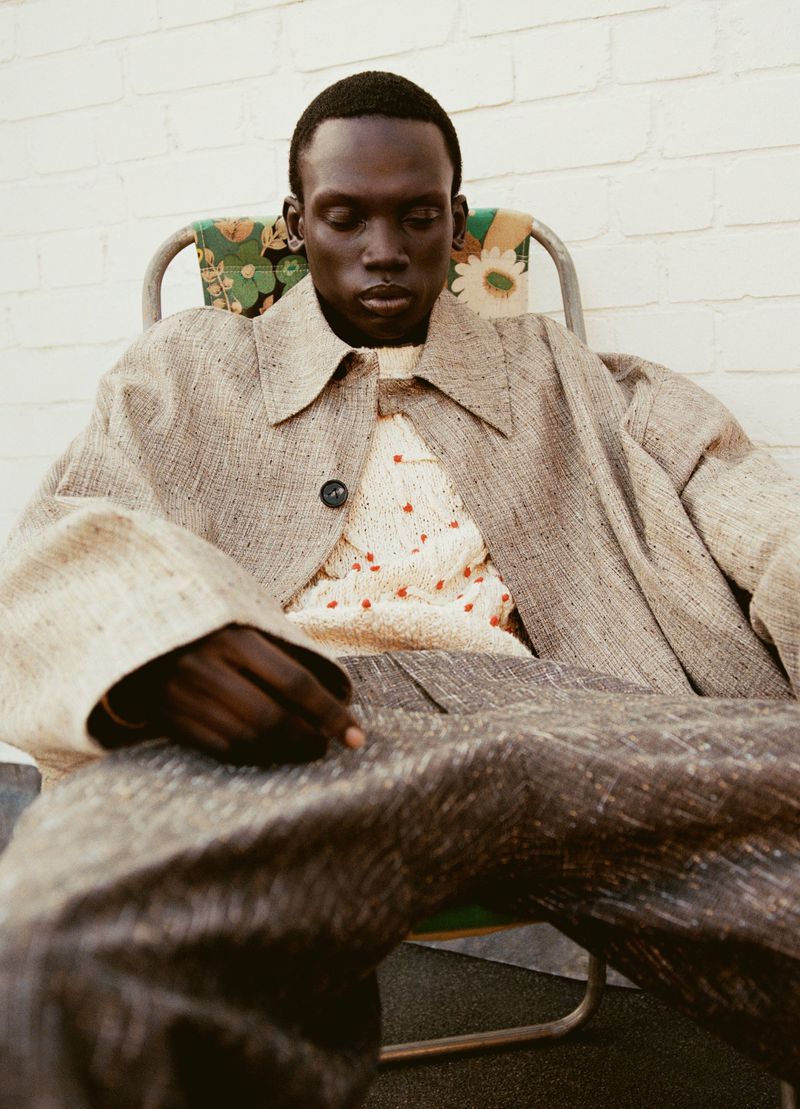 A pensive dark-skinned adult male model wearing a light coat and patterned sweater sits in a vintage lawn chair outdoors.