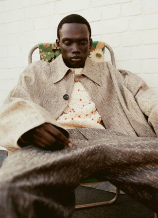 A pensive dark-skinned adult male model wearing a light coat and patterned sweater sits in a vintage lawn chair outdoors.