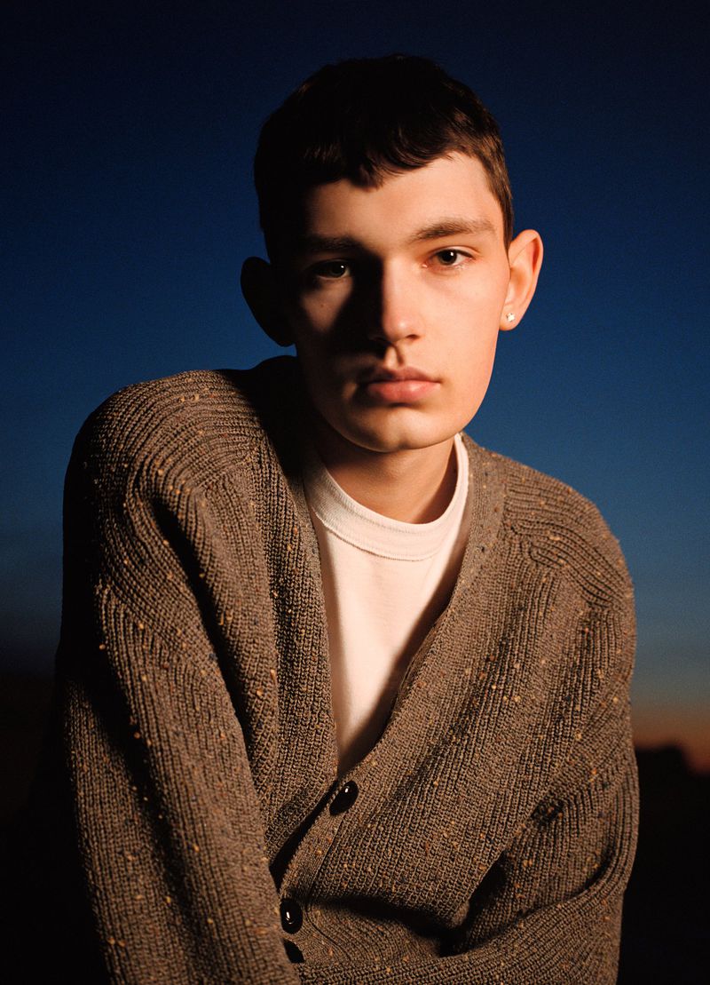 Portrait of a young adult male wearing a brown cardigan and white shirt, set against a dark blue and orange sky background.