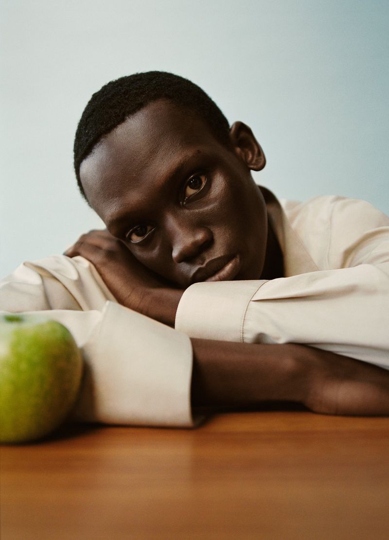 Portrait of a dark-skinned adult male resting his arms on a wooden table, with a green apple on the table next to him.