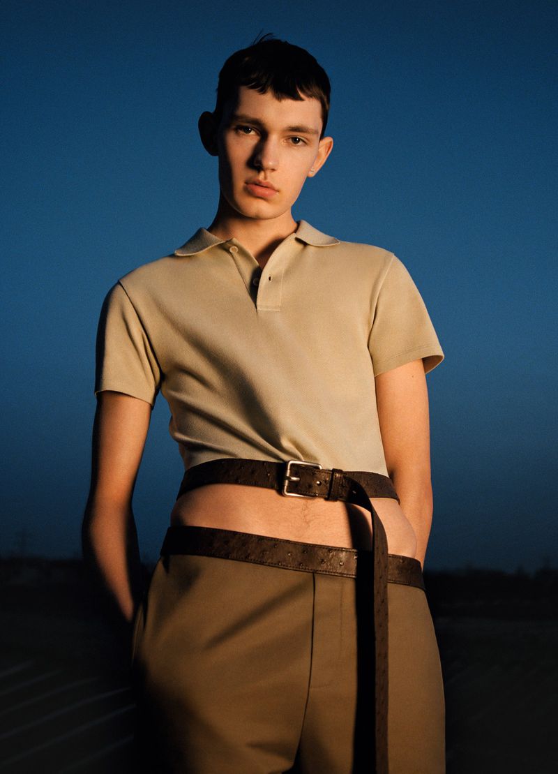 Portrait of a young adult male model in a beige polo shirt and brown pants with layered belts outdoors against a twilight-blue sky.