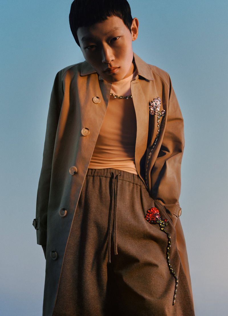 Asian woman in studio, posing in a tan trench coat with two flower brooches. She is wearing a peach long sleeve shirt and grey pants.