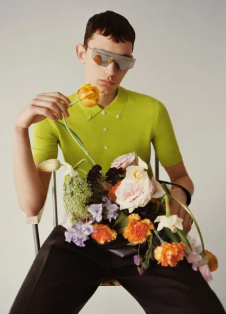 A stylish young adult sits holding flowers, wearing sunglasses, a chartreuse top, and dark pants against a neutral backdrop.