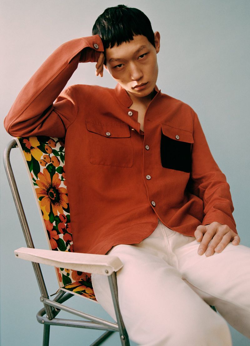 An Asian male model poses in an orange shirt and white pants while sitting on a floral-patterned chair in a light blue studio setting.