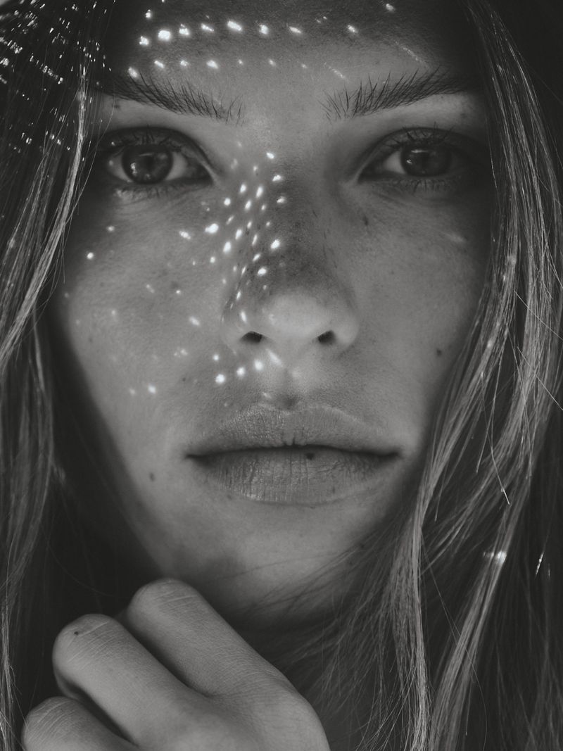 Close-up monochrome portrait of a woman with speckled light patterns on her face, long wavy hair, and hand near her chin.