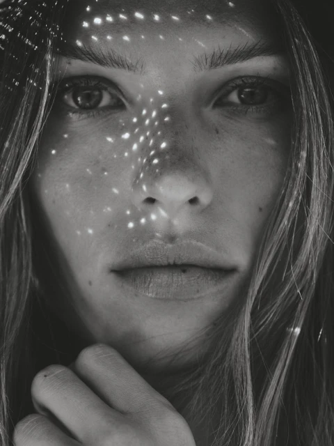 Close-up monochrome portrait of a woman with speckled light patterns on her face, long wavy hair, and hand near her chin.
