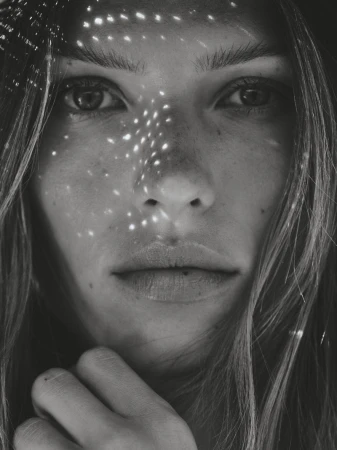 Close-up monochrome portrait of a woman with speckled light patterns on her face, long wavy hair, and hand near her chin.