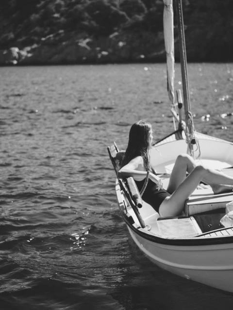 Monochrome image of a woman in a swimsuit lounging in a small boat on the water, with a forested coastline visible in the distance.