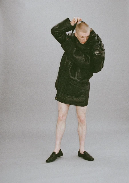 Adult male in a dark, puffy jacket with a backpack slung over his shoulder, standing in a photo studio against a grey backdrop.
