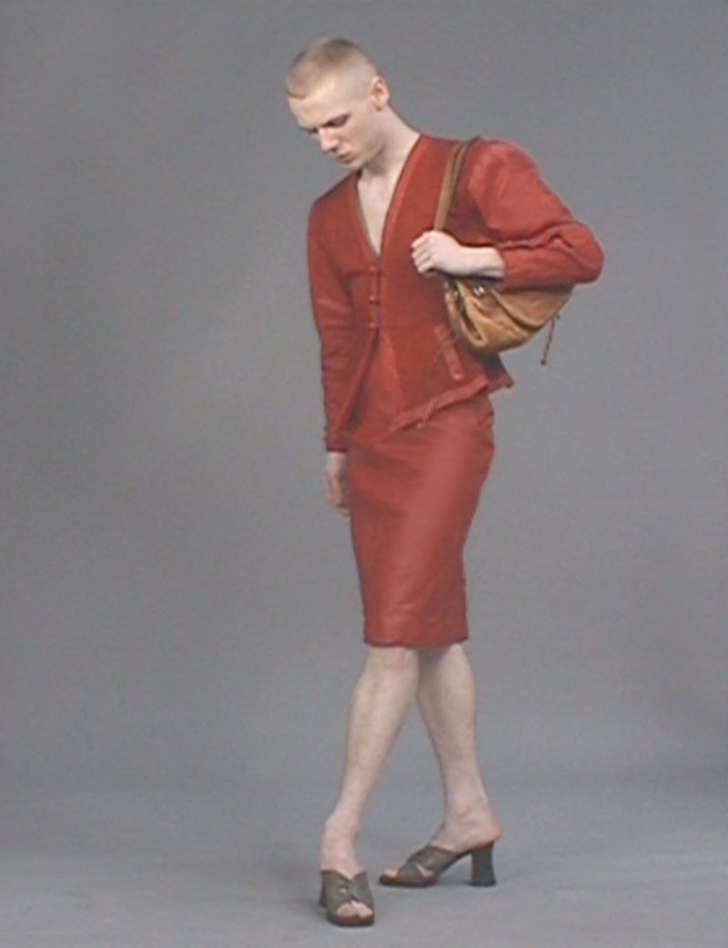 A blond adult wears a red suit with a handbag, posing in a studio with a neutral gray backdrop and wearing heels, looking down.