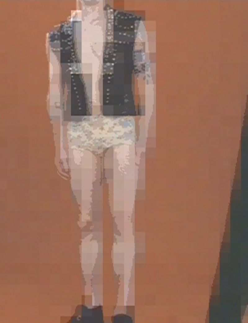 Full body shot of a person in an open studded vest, patterned shorts, and boots, against a solid backdrop.