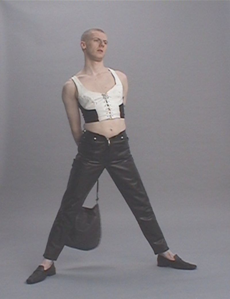 Non-binary fashion model strikes a pose in a studio wearing a cropped white and black top, dark pants, and loafers while carrying a handbag.