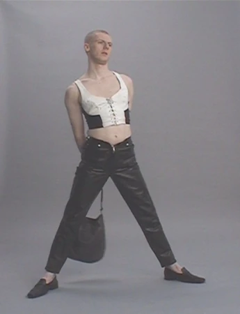 Non-binary fashion model strikes a pose in a studio wearing a cropped white and black top, dark pants, and loafers while carrying a handbag.
