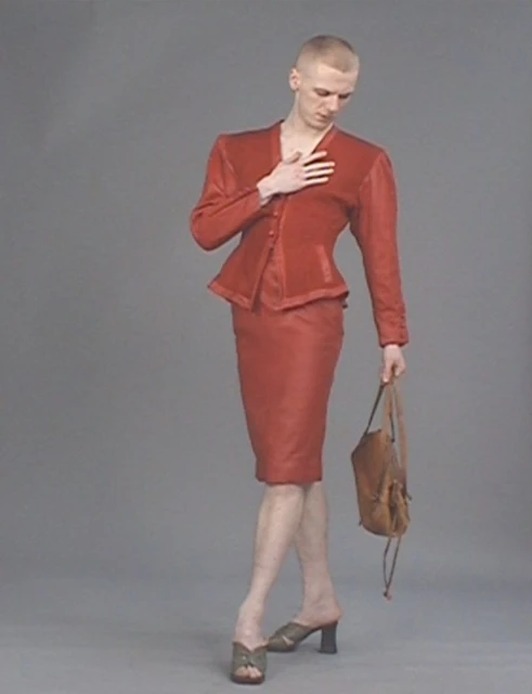 A slender person with a buzzcut poses in a reddish-brown skirt suit, holding a brown handbag against a neutral grey backdrop.