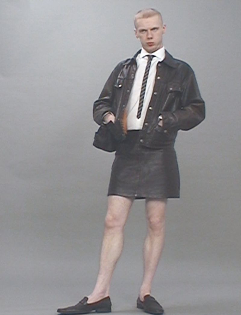 Young adult male with blond hair stands in leather skirt and jacket, white shirt, and tie, against gray backdrop in studio light.