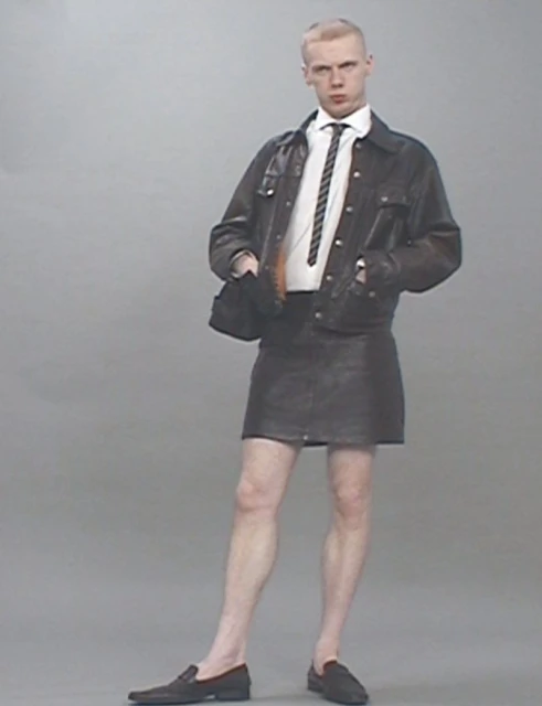 Young adult male with blond hair stands in leather skirt and jacket, white shirt, and tie, against gray backdrop in studio light.