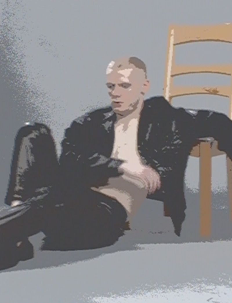 Man in a black leather jacket poses in front of a chair in a photo studio with gray background, showcasing a fashion and portrait style.