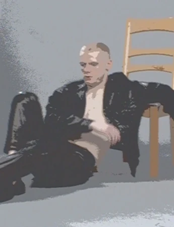 Man in a black leather jacket poses in front of a chair in a photo studio with gray background, showcasing a fashion and portrait style.