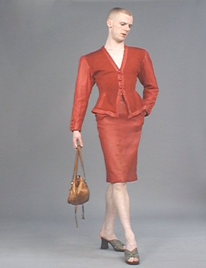 A person with a shaved head posing in a red skirt suit with a purse and heeled shoes in a studio setting with a plain gray background.