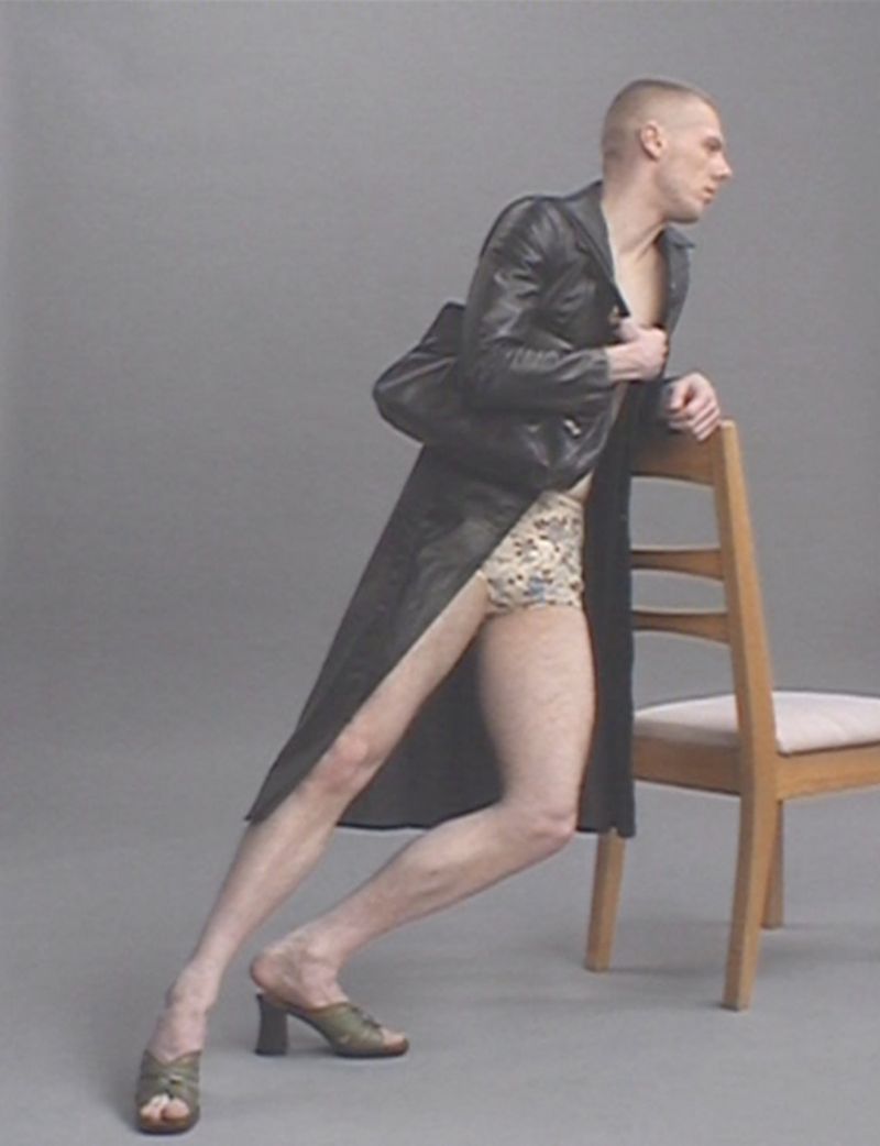 Stylish adult male poses with a wooden chair in a photo studio, wearing a leather trench coat and high-heeled mules.