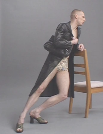 Stylish adult male poses with a wooden chair in a photo studio, wearing a leather trench coat and high-heeled mules.