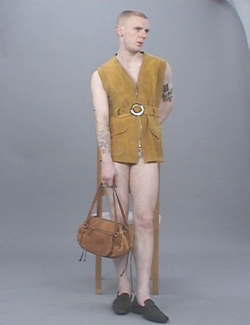 A fair-skinned adult male in a sleeveless gold vest and black shoes stands in a studio, holding a brown leather purse in his left hand.