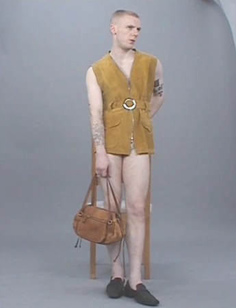 A fair-skinned adult male in a sleeveless gold vest and black shoes stands in a studio, holding a brown leather purse in his left hand.