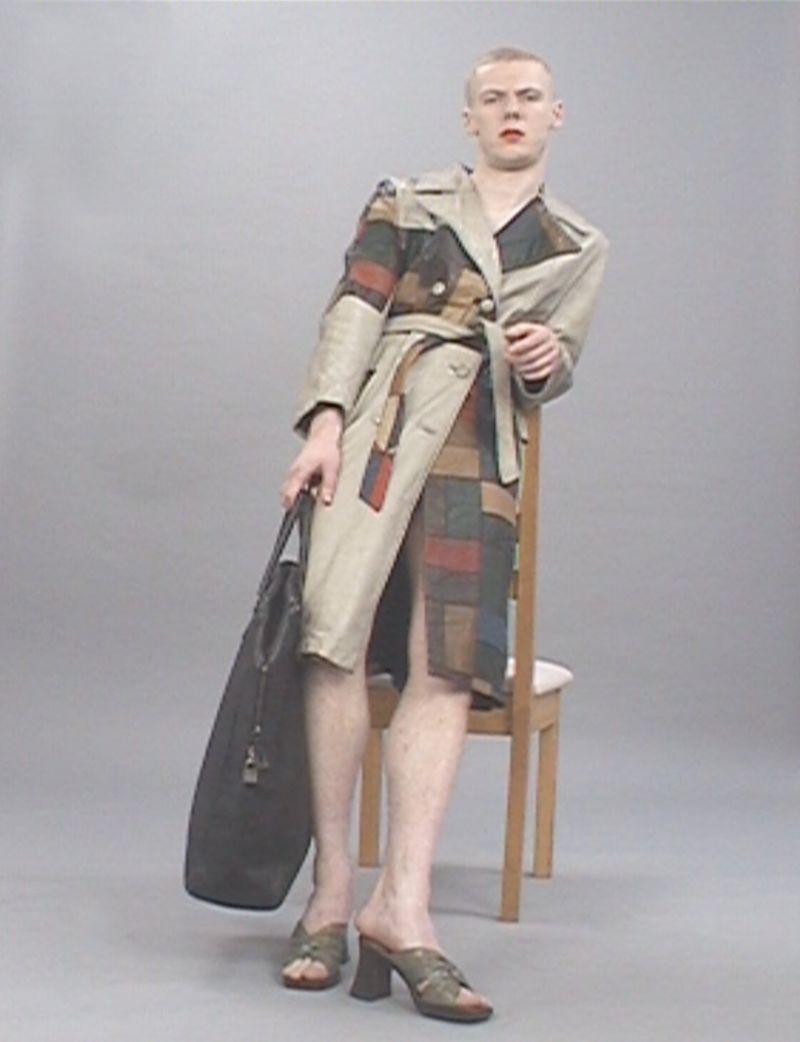 Man in a patchwork trench coat leans against a chair, holding a tote bag, posing in a neutral studio backdrop, showcasing an avant-garde fashion style.