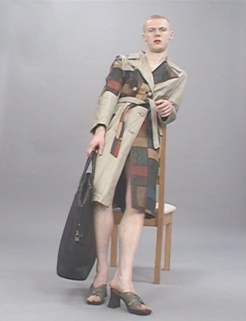 Man in a patchwork trench coat leans against a chair, holding a tote bag, posing in a neutral studio backdrop, showcasing an avant-garde fashion style.