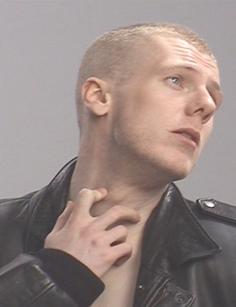 Close-up of an adult male with a shaved head wearing a black leather jacket, indoors, scratching his neck, looking upwards in studio lighting.
