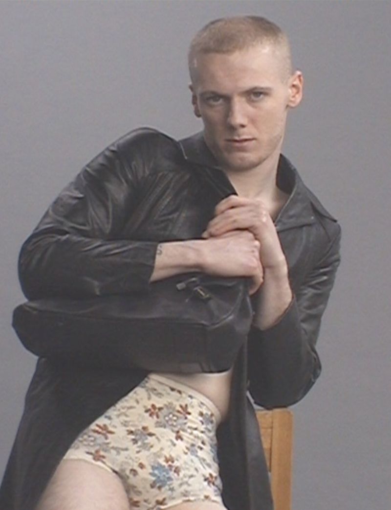 A person with short hair and a dark coat holds a bag, wearing floral patterned underwear, looking at the camera against a grey background.