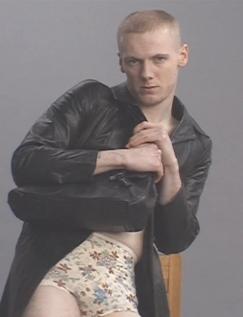 A person with short hair and a dark coat holds a bag, wearing floral patterned underwear, looking at the camera against a grey background.