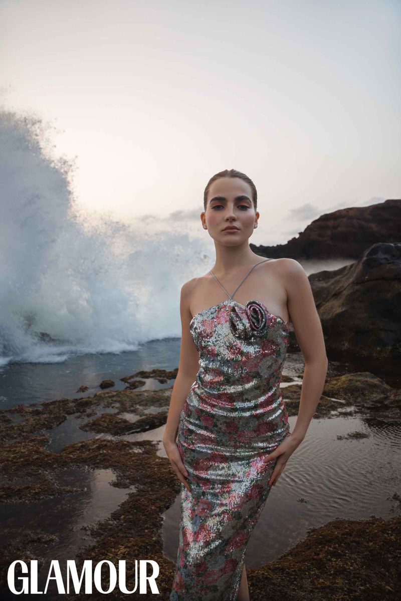 Glamour fashion editorial shot of a model in a floral dress posing on rocks with ocean waves crashing in the background during daytime.