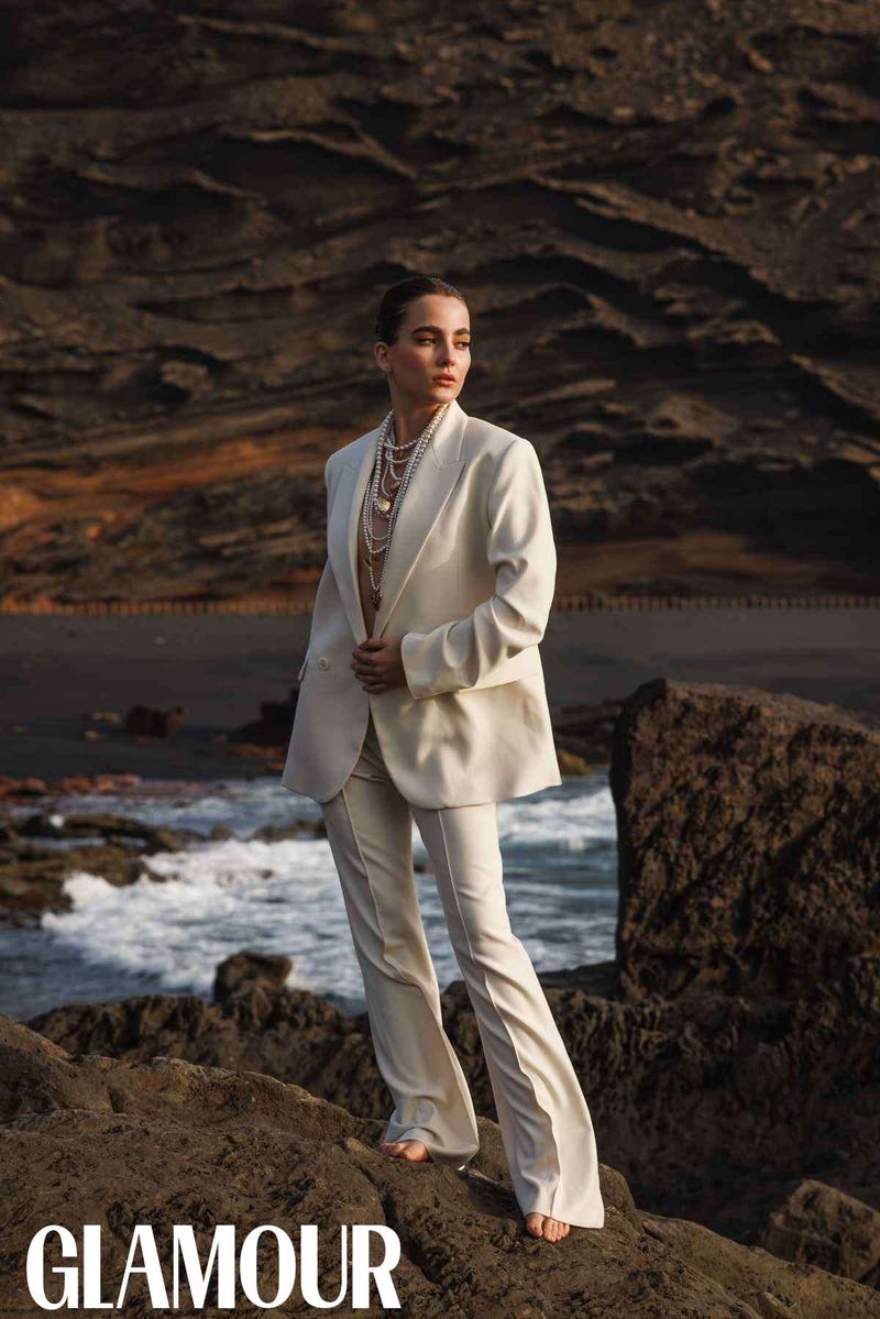 Woman in cream suit poses on rocky beach with ocean waves and dark cliffs, editorial fashion shot for Glamour magazine cover.